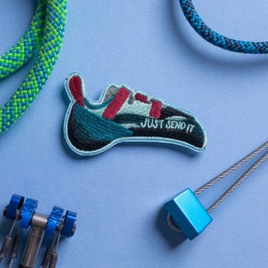 May include: A blue and green climbing shoe patch with the text "JUST SEND IT" in red thread. The patch is on a blue background with a blue and green rope and a blue carabiner.