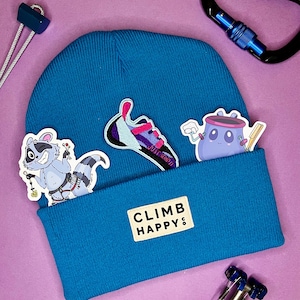 May include: A blue beanie with a white patch that says "CLIMB HAPPY" and three embroidered patches: a raccoon, a climbing shoe, and a purple pot with a face.
