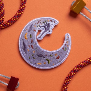 May include: A purple and white embroidered patch of a climber scaling a crescent moon-shaped rock face. The patch is surrounded by red and yellow rope.