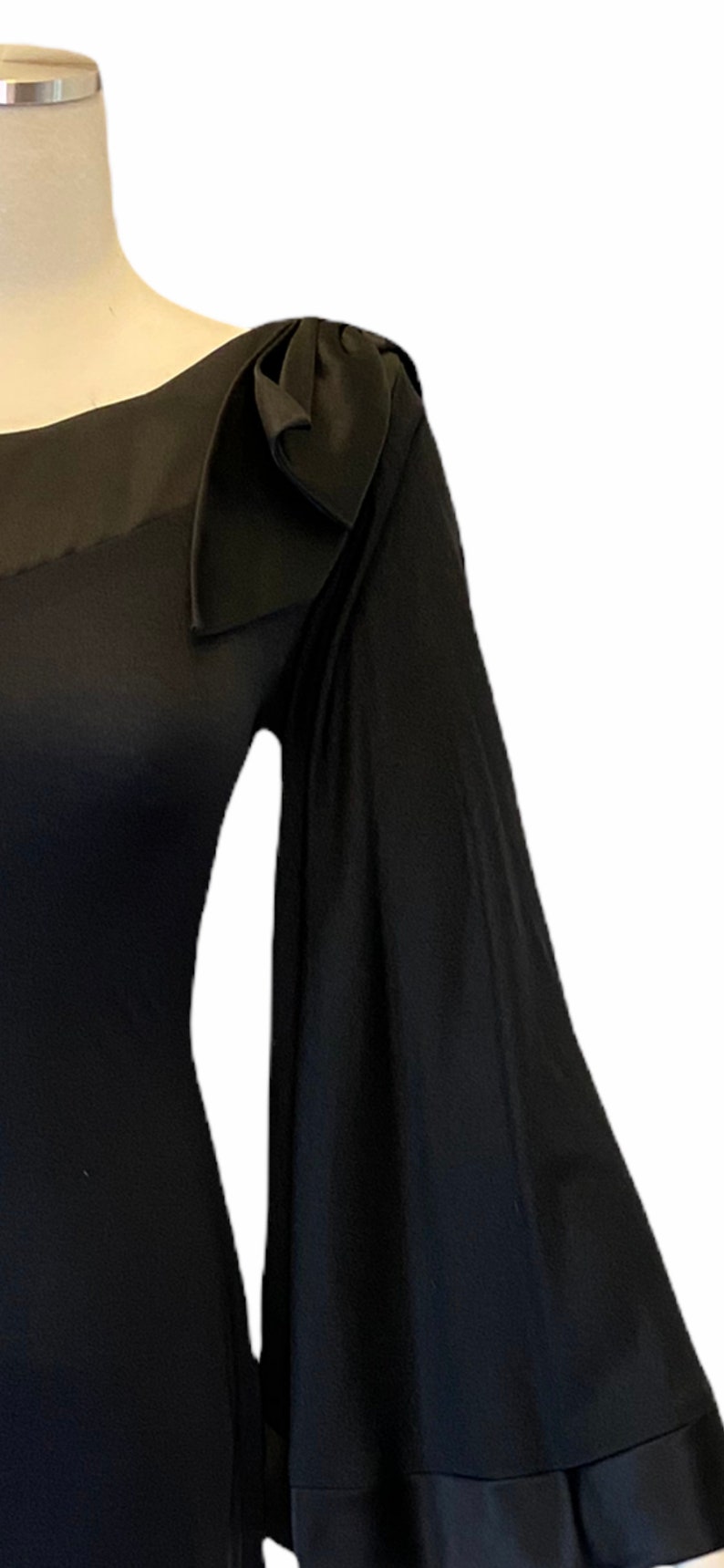 Puede incluir: Un vestido negro con un detalle de lazo &uacute;nico en el hombro. El vestido presenta una manga fluida en forma de campana. La tela parece tener textura, agregando profundidad. El vestido se exhibe en un maniqu&iacute;.