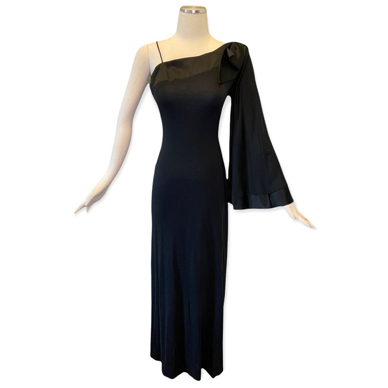 Puede incluir: Un vestido de noche negro de un solo hombro con una sola manga larga. El vestido presenta un detalle de lazo en el hombro y una silueta ajustada que llega hasta el suelo. La manga tiene un dise&ntilde;o acampanado con un ribete de sat&eacute;n.