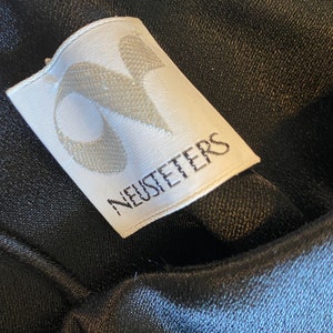 Puede incluir: Primer plano de una etiqueta de ropa blanca con la palabra "NEUSTETERS" impresa en negro. La etiqueta tiene un dise&ntilde;o estilizado en la esquina superior izquierda. La etiqueta est&aacute; adherida a una tela negra texturizada.