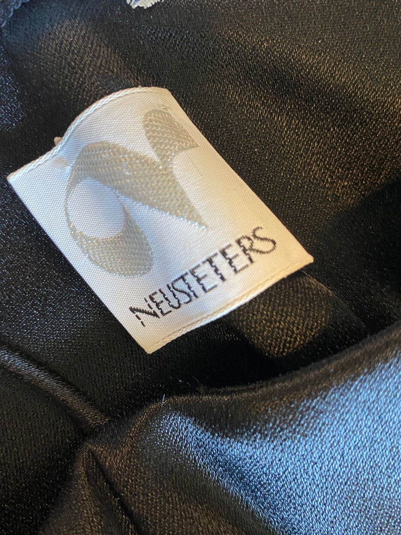 Puede incluir: Primer plano de una etiqueta de ropa blanca con la palabra "NEUSTETERS" impresa en negro. La etiqueta tiene un dise&ntilde;o estilizado en la esquina superior izquierda. La etiqueta est&aacute; adherida a una tela negra texturizada.
