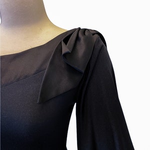 Puede incluir: Un vestido negro con escote cuadrado y mangas largas. El hombro presenta un detalle decorativo de lazo. El vestido se muestra en un maniqu&iacute; sobre un fondo blanco, destacando el dise&ntilde;o y la tela.