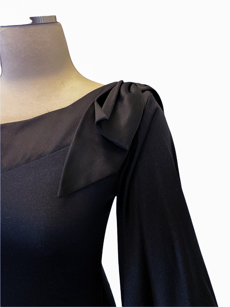 Puede incluir: Un vestido negro con escote cuadrado y mangas largas. El hombro presenta un detalle decorativo de lazo. El vestido se muestra en un maniqu&iacute; sobre un fondo blanco, destacando el dise&ntilde;o y la tela.