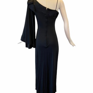 Puede incluir: Un vestido de noche negro de un solo hombro con una falda larga y fluida. El vestido presenta una manga larga con un pu&ntilde;o acampanado y una delicada correa de espagueti en el hombro opuesto. La parte posterior del vestido tiene un cierre de cremallera.