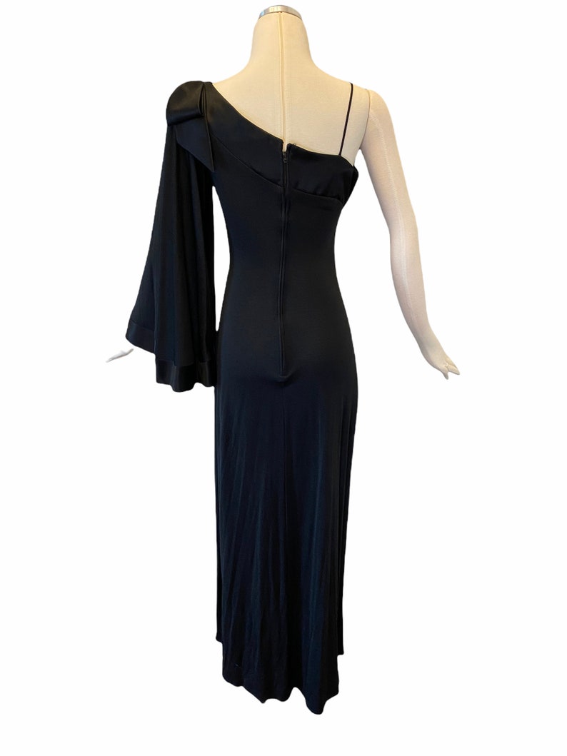 Puede incluir: Un vestido de noche negro de un solo hombro con una falda larga y fluida. El vestido presenta una manga larga con un pu&ntilde;o acampanado y una delicada correa de espagueti en el hombro opuesto. La parte posterior del vestido tiene un cierre de cremallera.