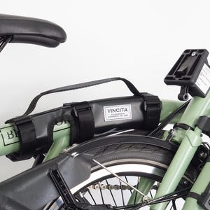 May include: A close-up of a folded green bicycle with black components. A black bag with the text "VINCITA" is attached to the frame. The bicycle has a black seat and tires.
