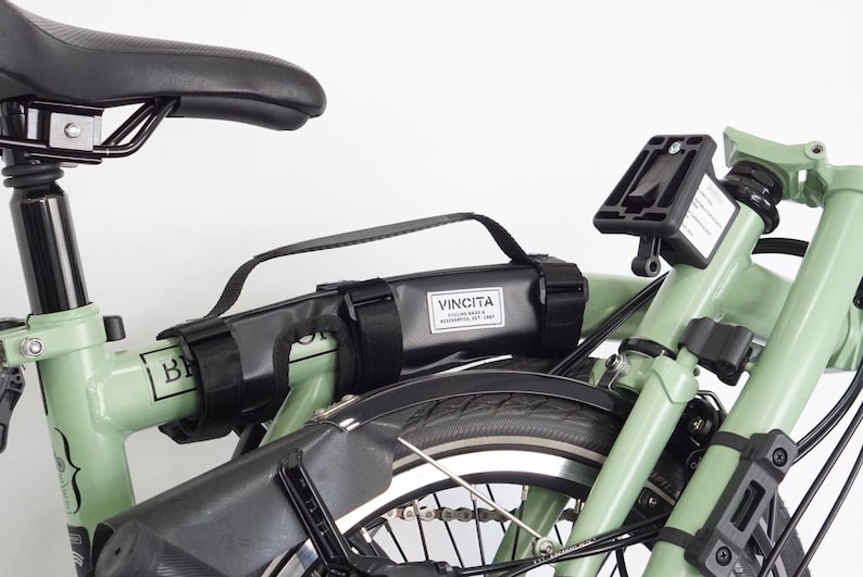 May include: A close-up of a folded green bicycle with black components. A black bag with the text "VINCITA" is attached to the frame. The bicycle has a black seat and tires.