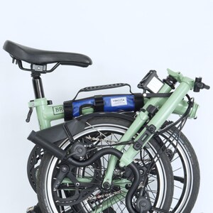 May include: A folded green bicycle with black tires and a black seat. A blue bag with the word "VINCITA" is attached to the frame. The bicycle is designed for compact storage and transport.