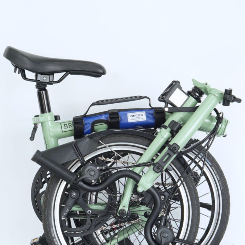 May include: A folded green bicycle with black tires and a black seat. A blue bag with the word "VINCITA" is attached to the frame. The bicycle is designed for compact storage and transport.