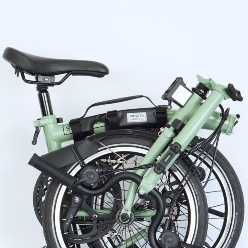 May include: Folded green folding bicycle with black tires, seat, and handlebars. A black bag with the word "VINCITA" is attached to the frame. The bicycle is designed for compact storage and transport.