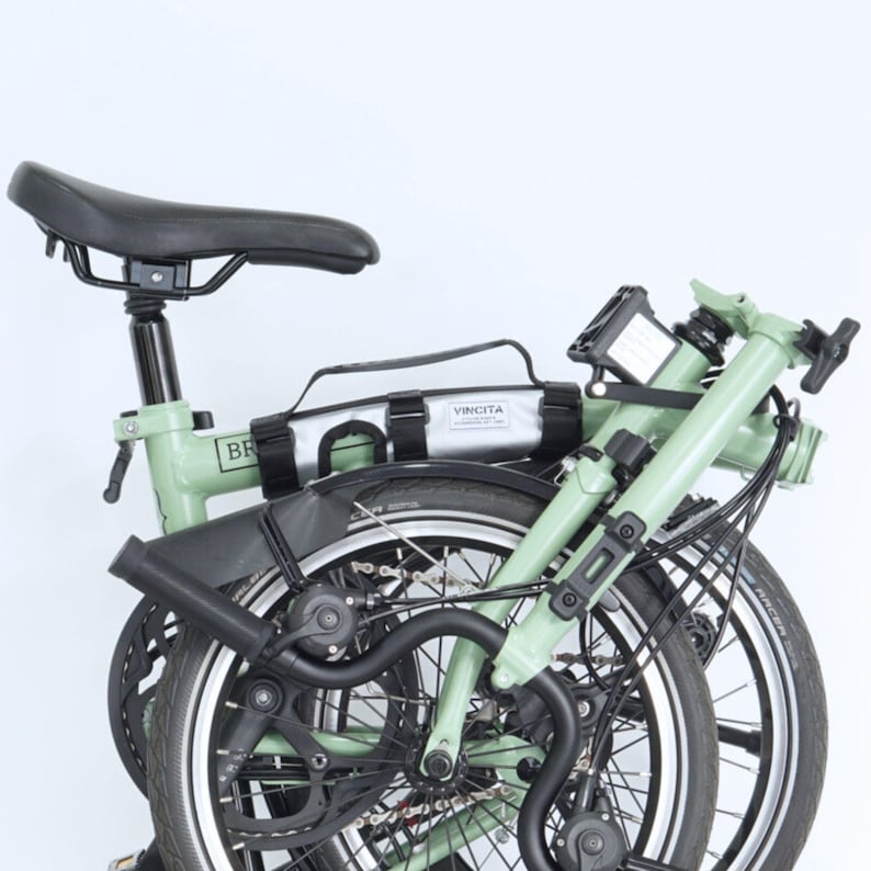 May include: A folded light green bicycle with black accents, including the seat, fenders, and tires. A silver bag with the text "VINCITA" is attached to the frame. The bicycle is set against a white background.