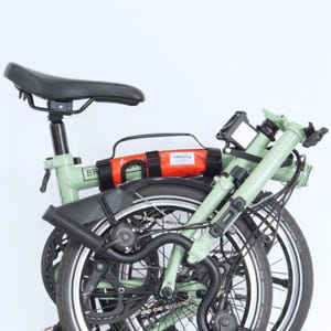 May include: A folded green bicycle with black tires and a black seat. An orange bag with the text "VINCITA" is attached to the frame. The bike is set against a white background.