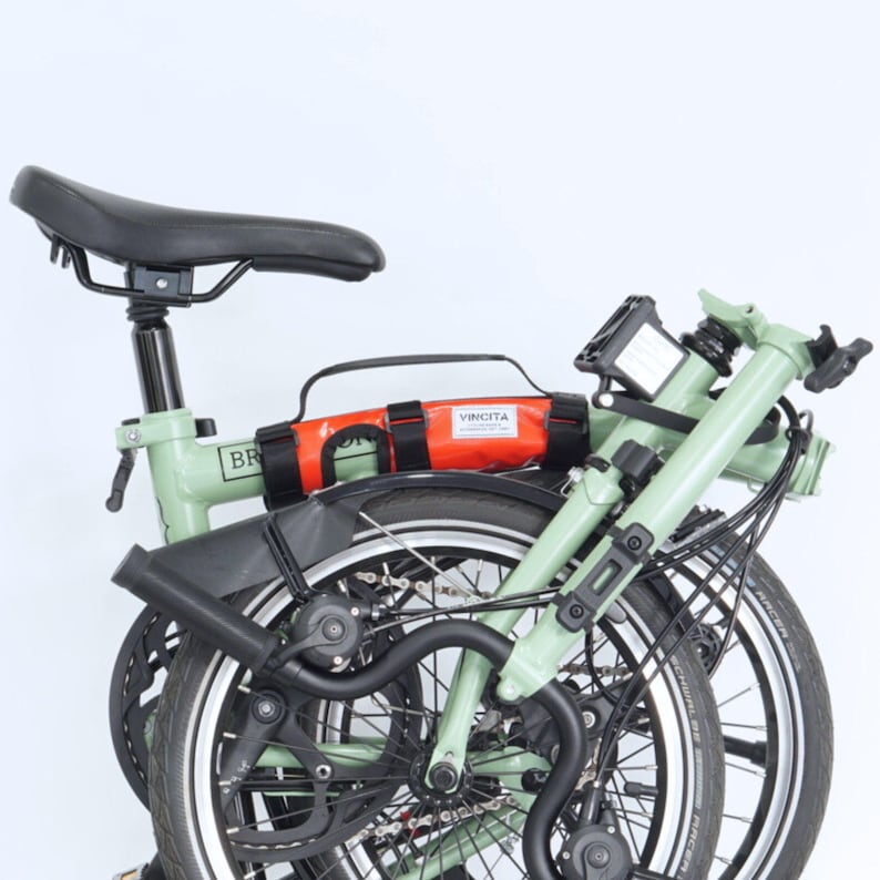 May include: A folded green bicycle with black tires and a black seat. An orange bag with the text "VINCITA" is attached to the frame. The bike is set against a white background.