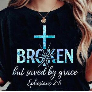 May include: A black t-shirt featuring a graphic with a blue and white watercolour cross and the word BROKEN, where the letter O is a shattered design. Below, the text reads but saved by grace and the scripture reference Ephesians 2:8.