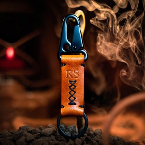 May include: A brown leather keychain with a black metal clasp and a key ring. The keychain is personalized with the initials "RS".