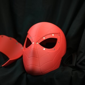 Puede incluir: Una m&aacute;scara de pl&aacute;stico roja con un dise&ntilde;o de telara&ntilde;a y agujeros para los ojos. La m&aacute;scara est&aacute; sobre una superficie negra.