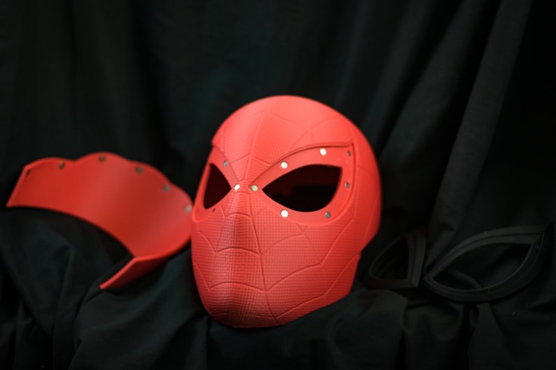 Puede incluir: Una m&aacute;scara de pl&aacute;stico roja con un dise&ntilde;o de telara&ntilde;a y agujeros para los ojos. La m&aacute;scara est&aacute; sobre una superficie negra.