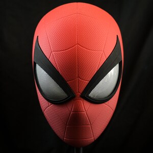 Puede incluir: Una m&aacute;scara de Spider-Man roja y negra con una superficie texturizada y un dise&ntilde;o de ojo de malla.
