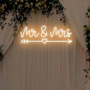 May include: White neon sign with the text "Mr & Mrs" and an arrow with a heart in the middle. The sign is hanging in front of a white curtain backdrop decorated with greenery.