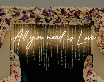 May include: A wedding ceremony arch decorated with pink and white flowers. The arch has a neon sign that reads "All you need is Love".