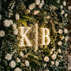 May include: A lush floral arrangement featuring white roses and greenery. A neon sign displays the letters "K" and "B" in a wedding or event setting. The backdrop is filled with various types of foliage.