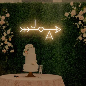 May include: A white wedding cake with pink flowers on top sits on a table with a pink lace tablecloth. A neon sign with the letters "J" and "A" and an arrow with a heart in the middle is on the wall behind the table.