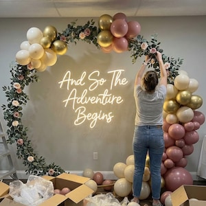 Puede incluir: Una mujer arreglando un arco de globos decorativo con el texto "And So The Adventure Begins". El arco presenta globos dorados, rosas y blancos, vegetación y flores. Cajas de cartón y una escalera están en primer plano.