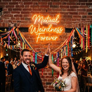 May include: A wedding scene with a neon sign saying "Mutual Weirdness Forever." A couple high-fives, celebrating. The brick wall is adorned with colourful ribbons and fairy lights. The bride holds a bouquet of flowers.