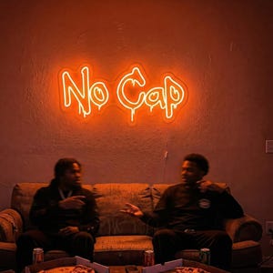 May include: An orange neon sign that reads "No Cap" hangs on a wall. Two people sit on a brown couch in front of a table with pizza boxes and pizza. The room is dimly lit with an orange glow.