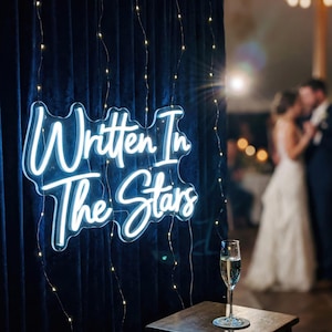 May include: A neon sign with the words "Written In The Stars" in white script is illuminated against a dark blue curtain. A glass of champagne sits on a small wooden table. Fairy lights hang from the curtain.