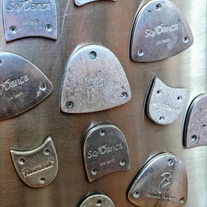 May include: A collection of silver-toned metal tap dance plates arranged on a metallic surface. Each plate features engraved brand names such as So Danca, Weissman, and Theatricals, along with small circular screw holes for attachment to footwear.