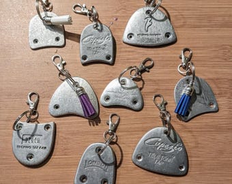 May include: A collection of silver-toned tap shoe keychains with various brand names, including Capezio and Bloch. Some keychains feature small, colorful tassels in white, purple, and blue. The keychains are displayed on a wooden surface.