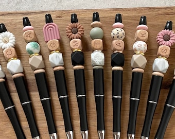 May include: Twelve black pens with different colored and patterned beads. The beads are made of wood, plastic, and stone. The pens are arranged in a row on a wooden surface.