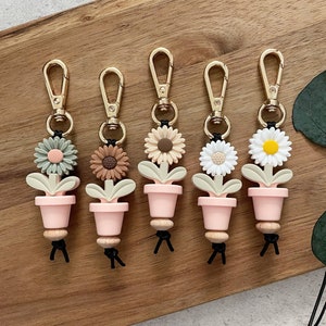 May include: Five keychains with gold rings and black cords. Each keychain features a pink pot with a green, brown, or white flower. The flowers are made of silicone and have a 3D effect.