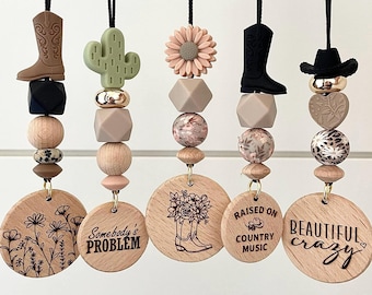 May include: Five wooden car charms with different designs and sayings. The charms are strung on black cords with various silicone beads. The designs include a cactus, a boot, a flower, a cowboy hat, and a boot with flowers. The sayings include "Somebody's Problem", "Raised on Country Music", and "Beautiful Crazy".