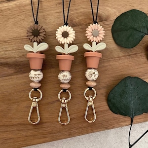May include: Three floral keychains with a brown, white, and pink flower charm, a brown pot charm, and a white bead with a floral design. Each keychain has a gold swivel clasp.
