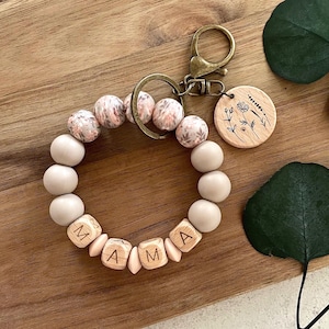 May include: A beaded keychain bracelet spelling "MAMA" with wooden letter beads. The bracelet has beige and patterned beads. A wooden charm with a floral design hangs from the clasp. The bracelet is on a wooden surface.