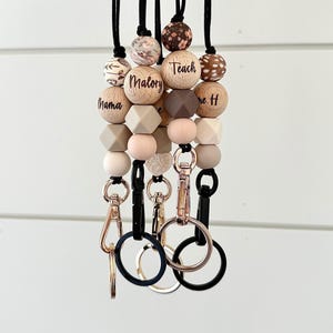 May include: Multiple beaded lanyards with black cords and metal clasps. The beads are in various shapes and colours, including wood, geometric, and round. Some beads have text, including "Mama", "Malory", and "Teach".