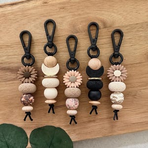 Könnte beinhalten: Fünf Schlüsselanhänger mit schwarzen Clips und dekorativen Perlen. Die Perlen umfassen Blumen-, Holz- und gemusterte Designs in Beige-, Rosa- und Schwarztönen. Jeder Schlüsselanhänger hat eine schwarze Quaste am unteren Ende. Die Schlüsselanhänger sind auf einer Holzoberfläche angeordnet.