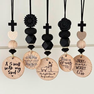 Peut inclure: Cinq breloques de voiture suspendues avec des pendentifs en forme de disque en bois. Chaque breloque comporte une croix noire, des perles et un disque en bois avec des citations et des illustrations inspirantes. Les breloques sont suspendues à des cordons noirs.