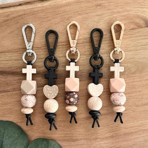 May include: Five keychains with various designs. Each keychain has a metal clasp, a cross-shaped bead, and a combination of wooden, patterned, and geometric beads in neutral tones. The keychains are arranged on a wooden surface.