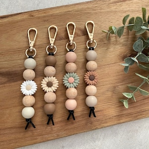 May include: Five wooden bead keychains with different coloured flower charms. The keychains are beige, brown, green, pink, and tan. Each keychain has a gold metal clasp.
