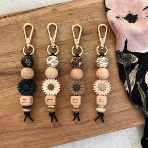 May include: Five wooden bead keychains with gold metal clasps. Each keychain has a different letter charm, a flower charm, and a patterned bead. The charms are in shades of brown, black, and pink.