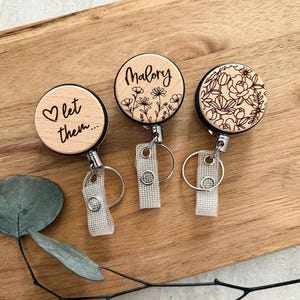 May include: Three wooden badge reels with different designs. One reel has the text "let them..." with a heart. Another has the name "Malory" with floral accents. The third reel features a floral pattern. Each reel has a silver clip and a clear strap.