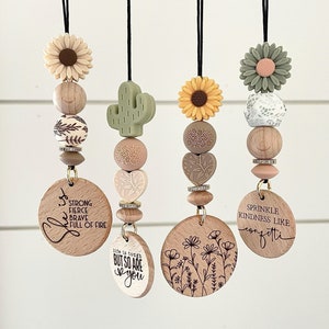 May include: Four wooden car charms with different designs. The charms are strung with beads and have a black cord for hanging. The designs include a sunflower, a cactus, a floral pattern, and a design with the text "Sprinkle Kindness Like Confetti".