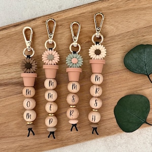 May include: Four keychains with gold-coloured clasps. Each keychain features wooden beads spelling "MOM", "MOM", "MOMS", and "MRSH". They are adorned with flower-shaped accents in various colours and small terracotta-coloured pots.