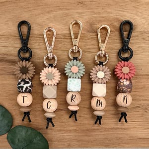 Könnte beinhalten: Fünf Schlüsselanhänger mit Gänseblümchen- und Perlenakzenten. Jeder Schlüsselanhänger hat ein anderes Farbschema, darunter schwarze, goldene und roségoldene Hardware. Die Perlen sind in verschiedenen Formen und Farben, mit Buchstaben auf einigen.