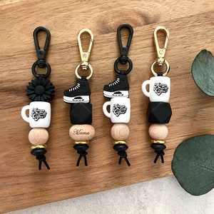 May include: Three black and white keychains with different charms. The charms include a coffee mug with the text "Mama Needs Coffee", a black ice skate, and a black flower. Each keychain has a gold clasp and black beads.